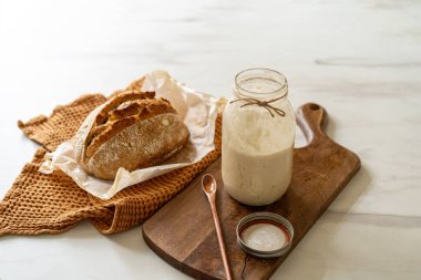 Çıtır ekşi ekmek, sıcak bir mutfak atmosferinde kırsal bir bez ve ahşap bir tahtanın eşlik ettiği etkin marş başlatıcısıyla dolu cam bir kavanozun yanında oturur..