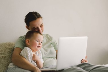 Evde dizüstü bilgisayar kullanan mutlu bir anne portresi. Birlikte, gülümseyen anne küçük oğluyla vakit geçirirken internette geziniyor..