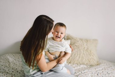 Evde çekilmiş bir erkek bebek portresi. Annesi yatakta dinleniyor ve hafta sonu birlikte yatakta oynuyor. Tembel bir sabah, sıcak ve samimi bir sahne. Anne çocuğunu kucağına alıp öpüyor. Anneler günü konsepti