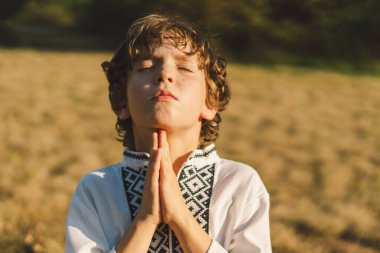 Çocuk, geleneksel işlemeli bir gömlek giyip, dua pozu vererek ayakta duruyor, ağaçların arasından süzülen yumuşak güneş ışığıyla sakin bir açık hava ortamının ortasında..