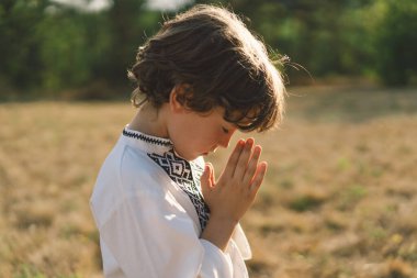 Çocuk, geleneksel işlemeli bir gömlek giyip, dua pozu vererek ayakta duruyor, ağaçların arasından süzülen yumuşak güneş ışığıyla sakin bir açık hava ortamının ortasında..