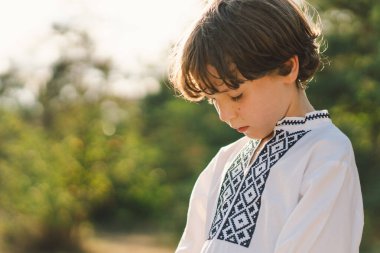Çocuk, geleneksel işlemeli bir gömlek giyip, dua pozu vererek ayakta duruyor, ağaçların arasından süzülen yumuşak güneş ışığıyla sakin bir açık hava ortamının ortasında..