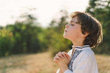 Çocuk, geleneksel işlemeli bir gömlek giyip, dua pozu vererek ayakta duruyor, ağaçların arasından süzülen yumuşak güneş ışığıyla sakin bir açık hava ortamının ortasında..