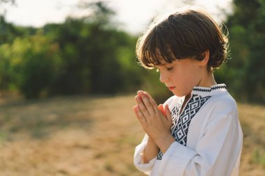 Çocuk, geleneksel işlemeli bir gömlek giyip, dua pozu vererek ayakta duruyor, ağaçların arasından süzülen yumuşak güneş ışığıyla sakin bir açık hava ortamının ortasında..