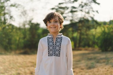 Genç bir çocuk, güneşli bir tarlada dikilirken özgüvenle gülümsüyor, öğleden sonra yemyeşil bir ortamda geleneksel işlemeli bir gömlek giyiyor.