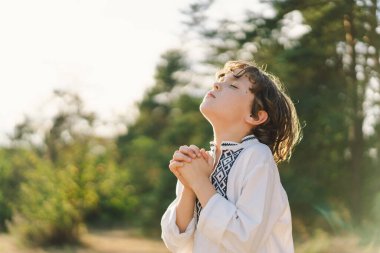 Çocuk, geleneksel işlemeli bir gömlek giyip, dua pozu vererek ayakta duruyor, ağaçların arasından süzülen yumuşak güneş ışığıyla sakin bir açık hava ortamının ortasında..