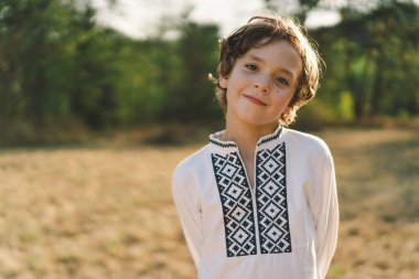 Genç bir çocuk, güneşli bir tarlada dikilirken özgüvenle gülümsüyor, öğleden sonra yemyeşil bir ortamda geleneksel işlemeli bir gömlek giyiyor.