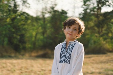 Genç bir çocuk, güneşli bir tarlada dikilirken özgüvenle gülümsüyor, öğleden sonra yemyeşil bir ortamda geleneksel işlemeli bir gömlek giyiyor.