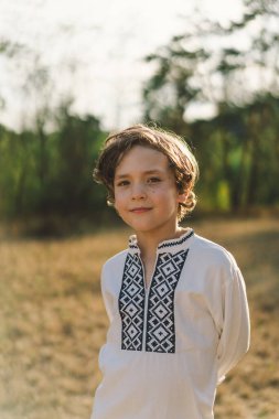 Genç bir çocuk, güneşli bir tarlada dikilirken özgüvenle gülümsüyor, öğleden sonra yemyeşil bir ortamda geleneksel işlemeli bir gömlek giyiyor.