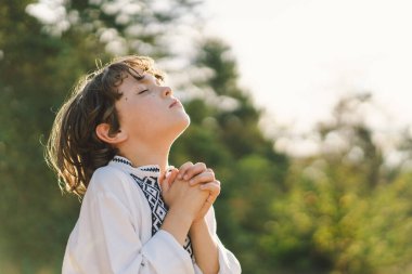 Çocuk, geleneksel işlemeli bir gömlek giyip, dua pozu vererek ayakta duruyor, ağaçların arasından süzülen yumuşak güneş ışığıyla sakin bir açık hava ortamının ortasında..