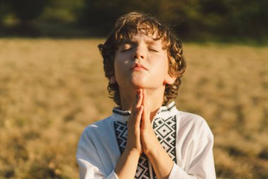 Çocuk, geleneksel işlemeli bir gömlek giyip, dua pozu vererek ayakta duruyor, ağaçların arasından süzülen yumuşak güneş ışığıyla sakin bir açık hava ortamının ortasında..