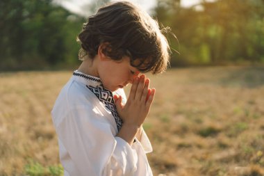 Çocuk, geleneksel işlemeli bir gömlek giyip, dua pozu vererek ayakta duruyor, ağaçların arasından süzülen yumuşak güneş ışığıyla sakin bir açık hava ortamının ortasında..