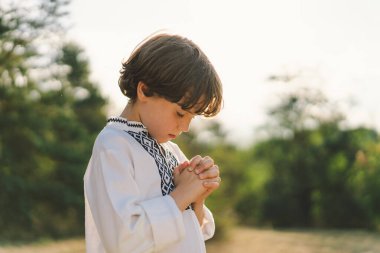 Çocuk, geleneksel işlemeli bir gömlek giyip, dua pozu vererek ayakta duruyor, ağaçların arasından süzülen yumuşak güneş ışığıyla sakin bir açık hava ortamının ortasında..