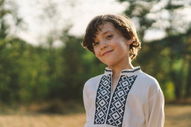 Genç bir çocuk, güneşli bir tarlada dikilirken özgüvenle gülümsüyor, öğleden sonra yemyeşil bir ortamda geleneksel işlemeli bir gömlek giyiyor.