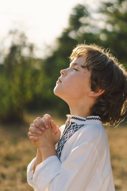 Çocuk, geleneksel işlemeli bir gömlek giyip, dua pozu vererek ayakta duruyor, ağaçların arasından süzülen yumuşak güneş ışığıyla sakin bir açık hava ortamının ortasında..