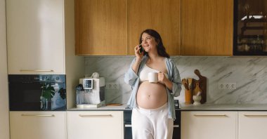 Hamile bir kadın şık bir mutfakta elinde fincanla telefonda konuşurken gülümsüyor. Alan ahşap dolaplar ve zarif aletler içeriyor, rahat bir atmosfer yaratıyor..