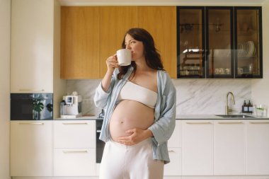 Hamile bir kadın şık bir mutfakta elinde bir fincan çayla duruyor ve huzurlu bir ortamda yaklaşan anneliği düşünüyor.