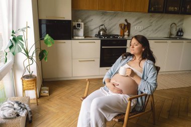 Sıcak bir mutfakta hamile bir kadın hamile karnını nazikçe tutuyor. Huzurlu bir ortamda yaklaşan anneliği yansıtıyor.