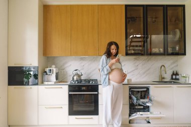 Hamile kadın çağdaş bir mutfakta duruyor, akıllı telefonuna odaklanmış. Doğal ışık mekanı aydınlatır, rahat ve şık bir ortam sunar..