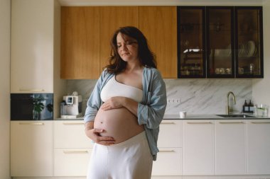 Sıcak bir mutfakta hamile bir kadın hamile karnını nazikçe tutuyor. Huzurlu bir ortamda yaklaşan anneliği yansıtıyor.