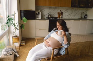 Sıcak bir mutfakta hamile bir kadın hamile karnını nazikçe tutuyor. Huzurlu bir ortamda yaklaşan anneliği yansıtıyor.