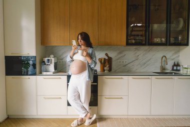 Hamile kadın çağdaş bir mutfakta duruyor, akıllı telefonuna odaklanmış. Doğal ışık mekanı aydınlatır, rahat ve şık bir ortam sunar..