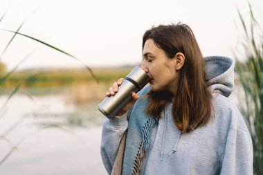 Nehir kenarında güzel bir gezgin kadın termostan sıcak bir içecek içiyor. Kadın turist nehir kenarındaki sonbahar manzarasının tadını çıkarıyor. Seyahat konsepti, rahatla..