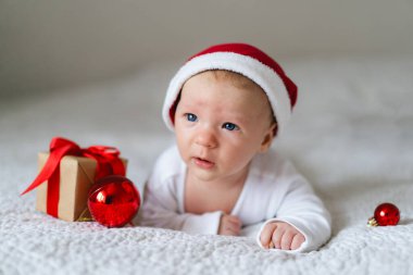 A baby wearing a red Santa hat lays on a cozy white blanket surrounded by festive decorations. A small gift box and shiny ornaments add to the cheerful holiday spirit.