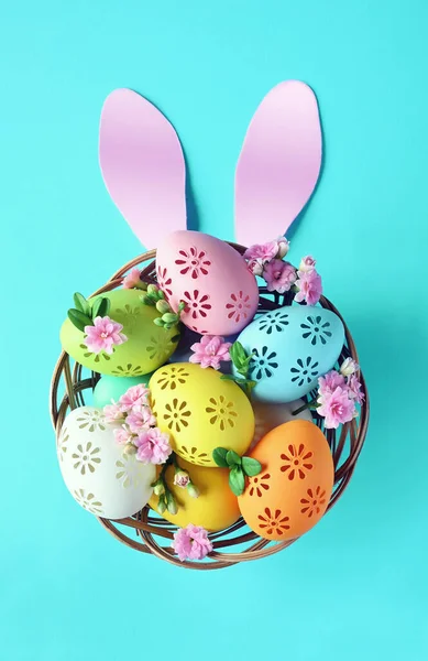 Multi colors Easter eggs in the basket with bunny ears and flowers on blue background. Pastel color Easter eggs