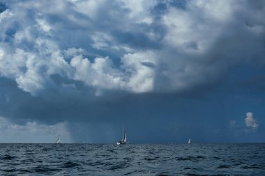 Ufukta dağılmış yelkenli yatlar açık denizde dramatik ve bulutlu bir gökyüzünün altında. Denizde dinamik dalgalar ve bot izleri var. Uzakta bir fırtına kopuyor. Yağmurdan önce yakalandı.