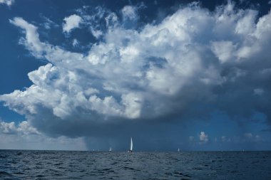 Ufukta dağılmış yelkenli yatlar açık denizde dramatik ve bulutlu bir gökyüzünün altında. Denizde dinamik dalgalar ve bot izleri var. Uzakta bir fırtına kopuyor. Yağmurdan önce yakalandı.
