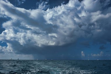 Ufukta dağılmış yelkenli yatlar açık denizde dramatik ve bulutlu bir gökyüzünün altında. Denizde dinamik dalgalar ve bot izleri var. Uzakta bir fırtına kopuyor. Yağmurdan önce yakalandı.