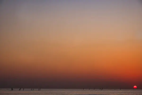 Sakin bir denizin üzerinde canlı turuncu ve kırmızı tonlarla minimum gün batımı manzarası. Güneş ufka yakın küçük kırmızı bir daire gibi görünür. Barışçıl ve soyut doğal sahne, arka plan veya meditasyon temaları için mükemmel.