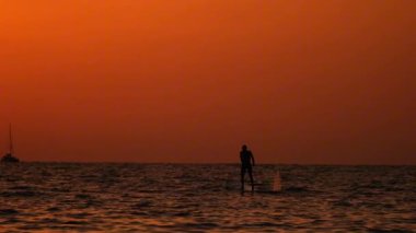 Parlayan turuncu bir günbatımında tek başına denizde kürek çeken bir adamın sinematik videosu. Ufukta uzak bir yat görünüyor. Huzurlu ve atmosferik deniz manzarası, seyahat, özgürlük ve yaşam tarzı temaları için mükemmel..