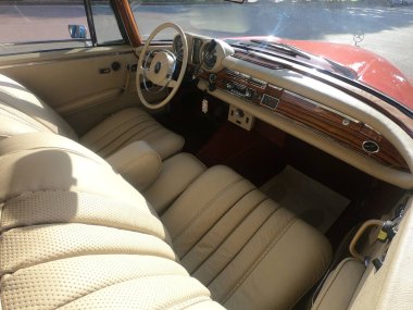 Caddeye güneş ışığı altında park edilmiş klasik kırmızı bir Mercedes Benz 280 SE Coupe. İkonik Alman lüks aracında krom bir ızgara, belirgin yuvarlak farlar ve 1960 'ların klasik tasarım detayları yer almaktadır..