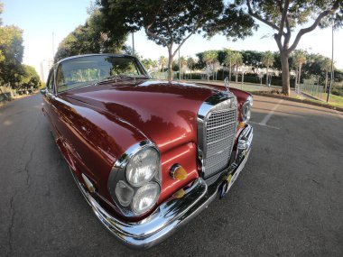 Caddeye güneş ışığı altında park edilmiş klasik kırmızı bir Mercedes Benz 280 SE Coupe. İkonik Alman lüks aracında krom bir ızgara, belirgin yuvarlak farlar ve 1960 'ların klasik tasarım detayları yer almaktadır..