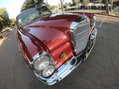 Caddeye güneş ışığı altında park edilmiş klasik kırmızı bir Mercedes Benz 280 SE Coupe. İkonik Alman lüks aracında krom bir ızgara, belirgin yuvarlak farlar ve 1960 'ların klasik tasarım detayları yer almaktadır..