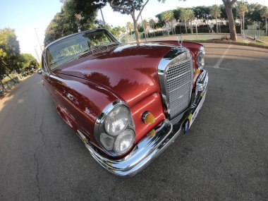 Caddeye güneş ışığı altında park edilmiş klasik kırmızı bir Mercedes Benz 280 SE Coupe. İkonik Alman lüks aracında krom bir ızgara, belirgin yuvarlak farlar ve 1960 'ların klasik tasarım detayları yer almaktadır..