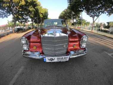 Caddeye güneş ışığı altında park edilmiş klasik kırmızı bir Mercedes Benz 280 SE Coupe. İkonik Alman lüks aracında krom bir ızgara, belirgin yuvarlak farlar ve 1960 'ların klasik tasarım detayları yer almaktadır..