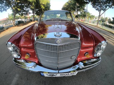 Caddeye güneş ışığı altında park edilmiş klasik kırmızı bir Mercedes Benz 280 SE Coupe. İkonik Alman lüks aracında krom bir ızgara, belirgin yuvarlak farlar ve 1960 'ların klasik tasarım detayları yer almaktadır..