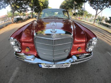 Caddeye güneş ışığı altında park edilmiş klasik kırmızı bir Mercedes Benz 280 SE Coupe. İkonik Alman lüks aracında krom bir ızgara, belirgin yuvarlak farlar ve 1960 'ların klasik tasarım detayları yer almaktadır..