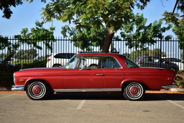 Caddeye güneş ışığı altında park edilmiş klasik kırmızı bir Mercedes Benz 280 SE Coupe. İkonik Alman lüks aracında krom bir ızgara, belirgin yuvarlak farlar ve 1960 'ların klasik tasarım detayları yer almaktadır..