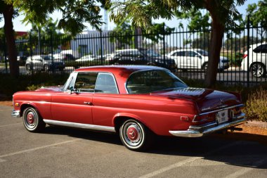 Caddeye güneş ışığı altında park edilmiş klasik kırmızı bir Mercedes Benz 280 SE Coupe. İkonik Alman lüks aracında krom bir ızgara, belirgin yuvarlak farlar ve 1960 'ların klasik tasarım detayları yer almaktadır..