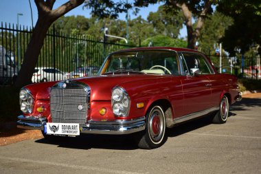 Caddeye güneş ışığı altında park edilmiş klasik kırmızı bir Mercedes Benz 280 SE Coupe. İkonik Alman lüks aracında krom bir ızgara, belirgin yuvarlak farlar ve 1960 'ların klasik tasarım detayları yer almaktadır..