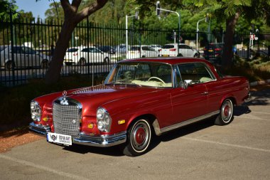 Caddeye güneş ışığı altında park edilmiş klasik kırmızı bir Mercedes Benz 280 SE Coupe. İkonik Alman lüks aracında krom bir ızgara, belirgin yuvarlak farlar ve 1960 'ların klasik tasarım detayları yer almaktadır..