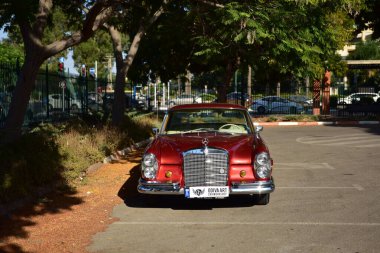 Caddeye güneş ışığı altında park edilmiş klasik kırmızı bir Mercedes Benz 280 SE Coupe. İkonik Alman lüks aracında krom bir ızgara, belirgin yuvarlak farlar ve 1960 'ların klasik tasarım detayları yer almaktadır..