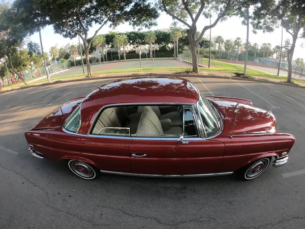 Caddeye güneş ışığı altında park edilmiş klasik kırmızı bir Mercedes Benz 280 SE Coupe. İkonik Alman lüks aracında krom bir ızgara, belirgin yuvarlak farlar ve 1960 'ların klasik tasarım detayları yer almaktadır..