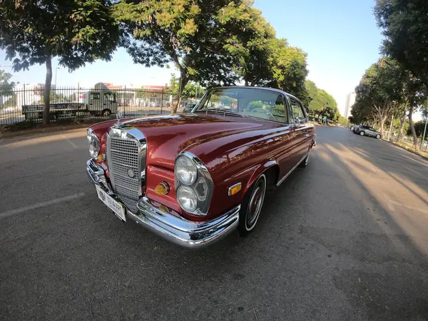 Caddeye güneş ışığı altında park edilmiş klasik kırmızı bir Mercedes Benz 280 SE Coupe. İkonik Alman lüks aracında krom bir ızgara, belirgin yuvarlak farlar ve 1960 'ların klasik tasarım detayları yer almaktadır..