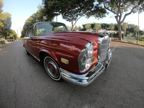 Caddeye güneş ışığı altında park edilmiş klasik kırmızı bir Mercedes Benz 280 SE Coupe. İkonik Alman lüks aracında krom bir ızgara, belirgin yuvarlak farlar ve 1960 'ların klasik tasarım detayları yer almaktadır..