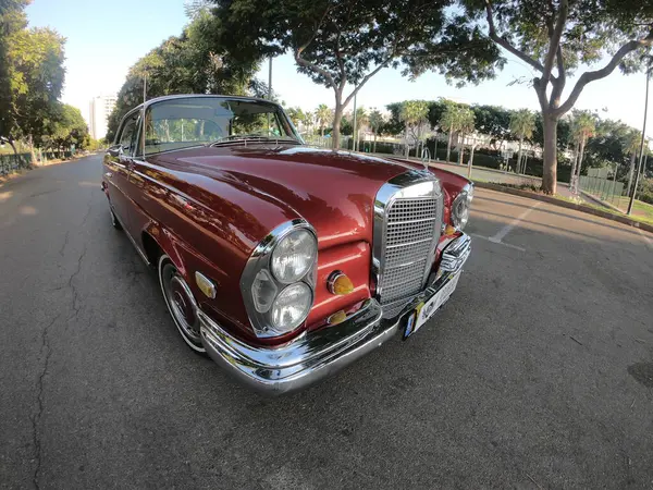 Caddeye güneş ışığı altında park edilmiş klasik kırmızı bir Mercedes Benz 280 SE Coupe. İkonik Alman lüks aracında krom bir ızgara, belirgin yuvarlak farlar ve 1960 'ların klasik tasarım detayları yer almaktadır..